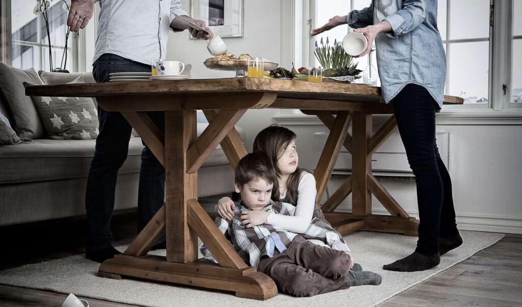 Children huddle under a wooden dining table as adults prepare breakfast nearby, creating a tense, hiding moment in a bright kitchen.”] ,