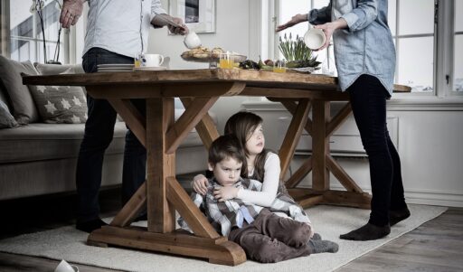 Children huddle under a wooden dining table as adults prepare breakfast nearby, creating a tense, hiding moment in a bright kitchen.”] ,